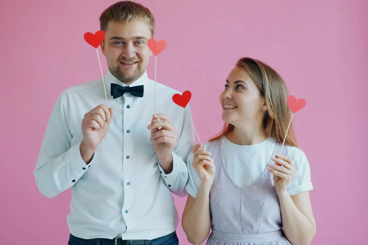 A couple holding heart props on sticks in front of a pink backdrop